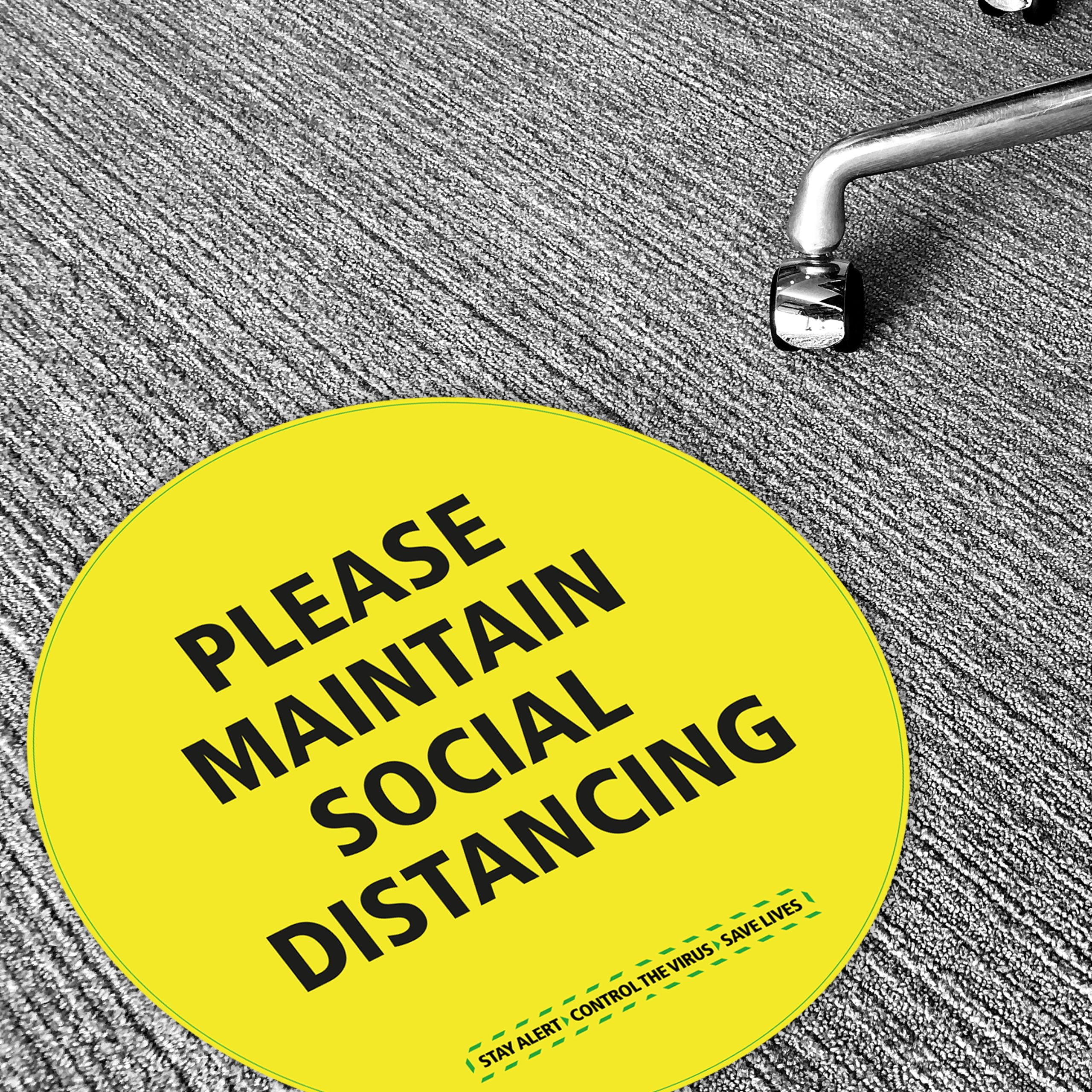 Yellow circular floor sticker reading “Please Maintain Social Distancing” applied to grey short pile carpet with castor chair in view